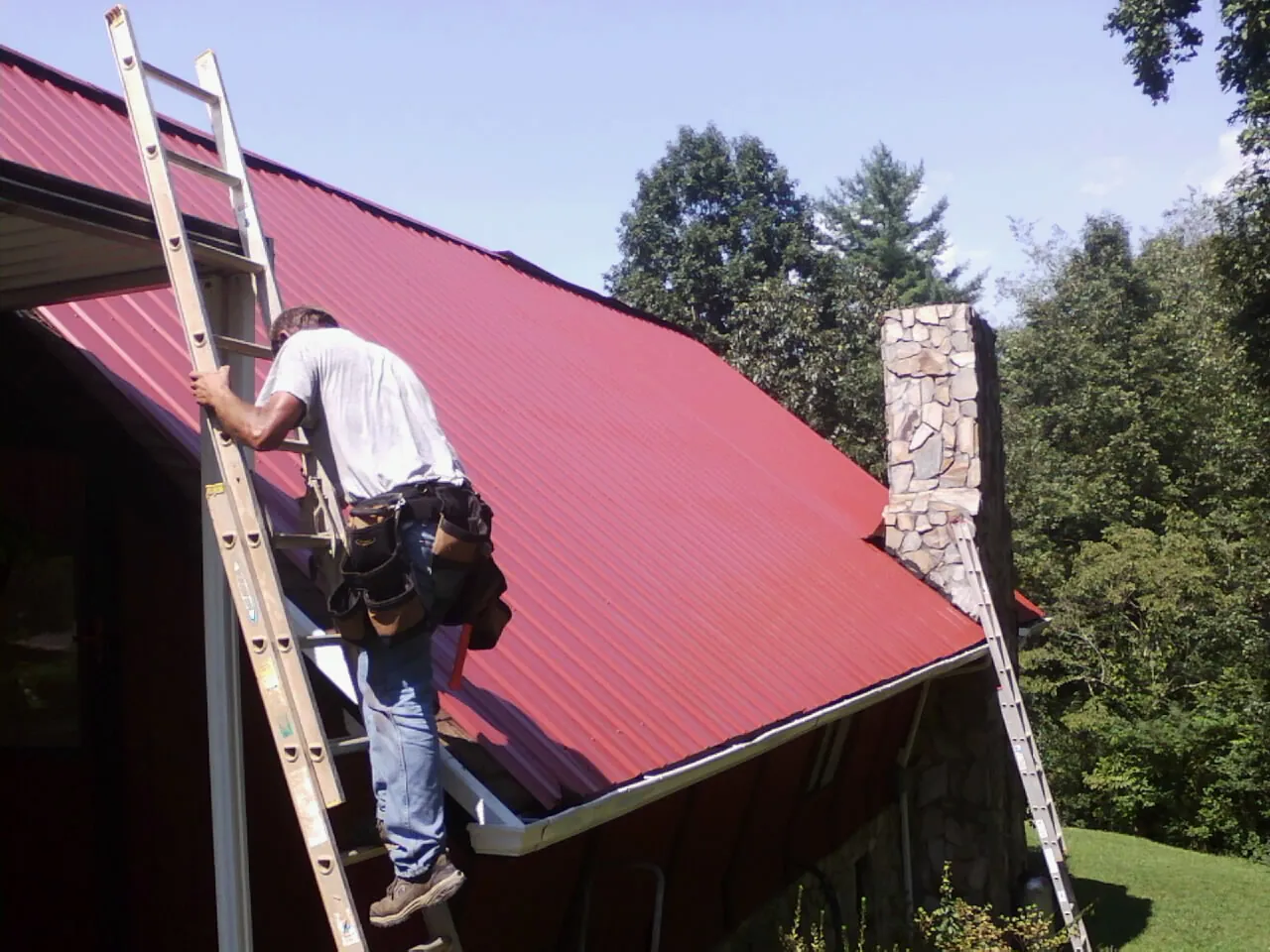 Roofer on a ladder beside a red metal roof and stone chimney — Soffit & Fascia Repair by Roofing Brothers in Craigmont